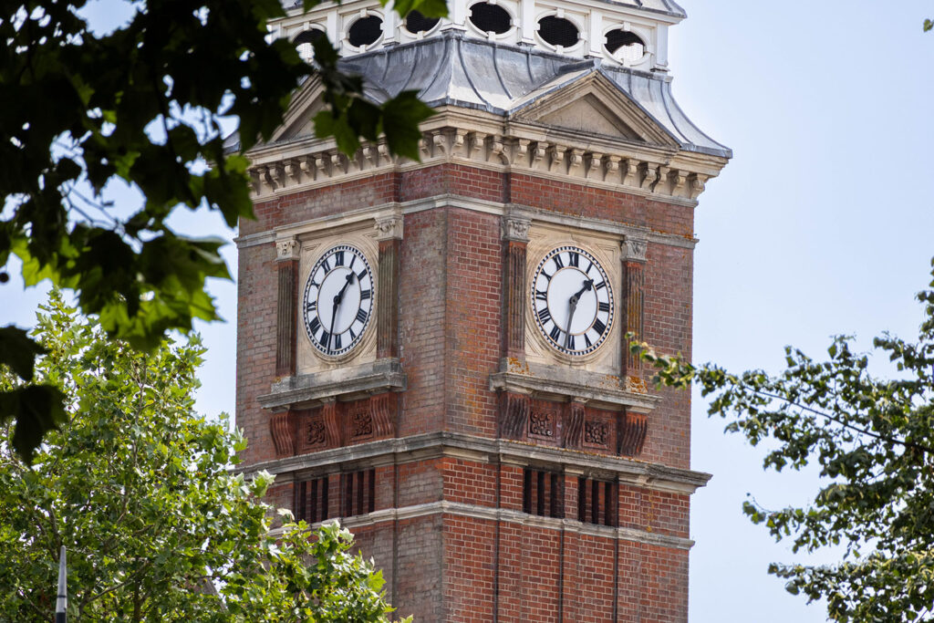 Picture of a clocktower with trees around the outside. The sky is quite pale.