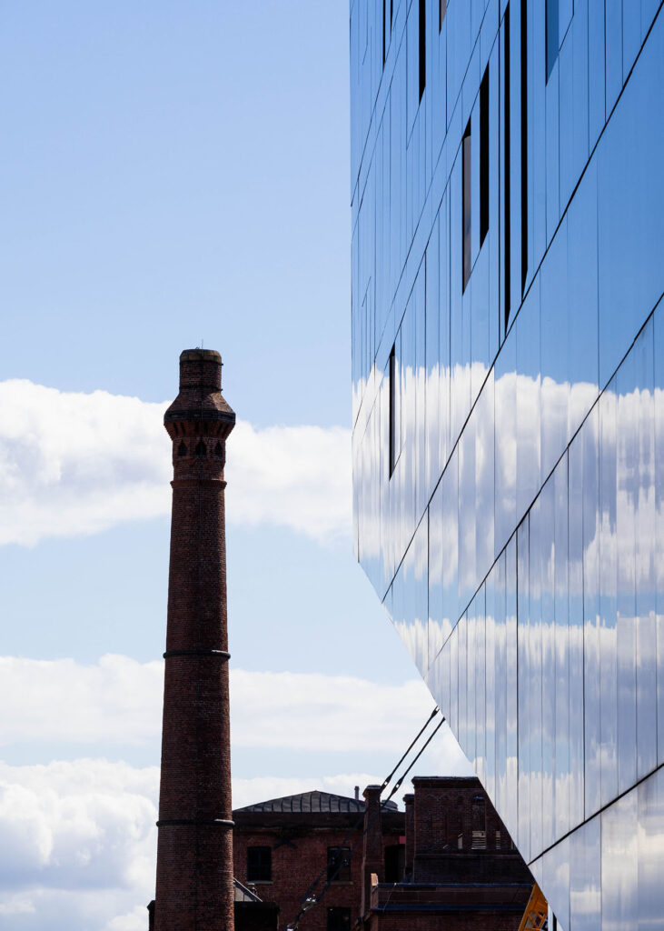 A picture of a pub with a smokestack, with a oddly shaped glass building in the foreground