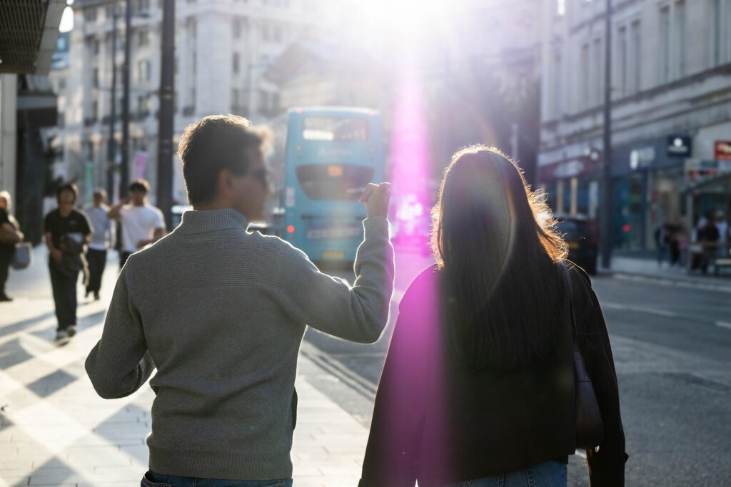 A picture of a man walking with a woman, with the man seemingly pointing towards the camera (face has been blurred for privacy). There is lens flare to an extent in the image, with it being visible on the female's hair and jacket.